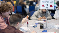 Students seated at a table pick up paperclips using an electromagnet made from a large nail, copper wire and a standard battery.
