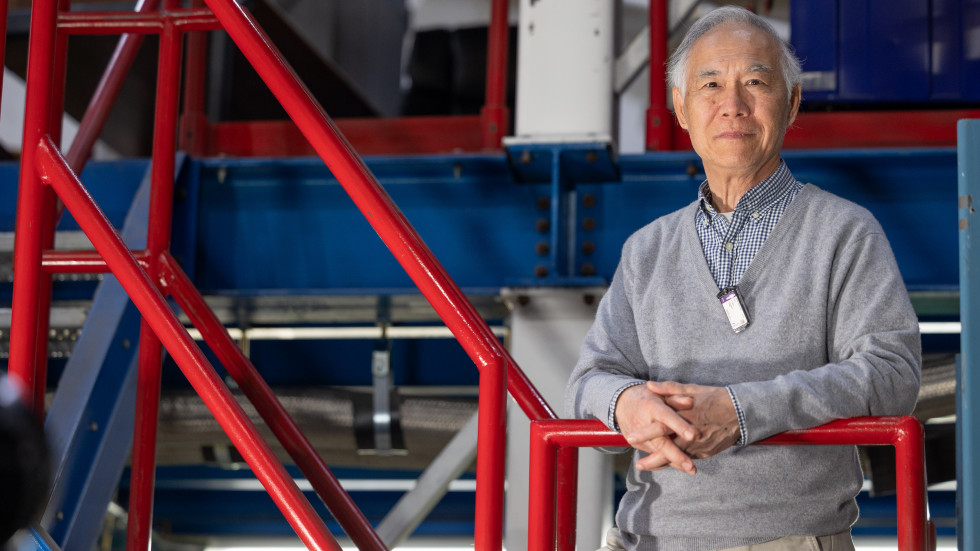 Image of Liguang Tang, Senior Staff Scientist & Physics Professor, standing in Experimental Hall C at Jefferson Lab with a confident expression. A quote overlays the image, emphasizing the importance of university resources and experiential learning in developing future thinkers and doers.