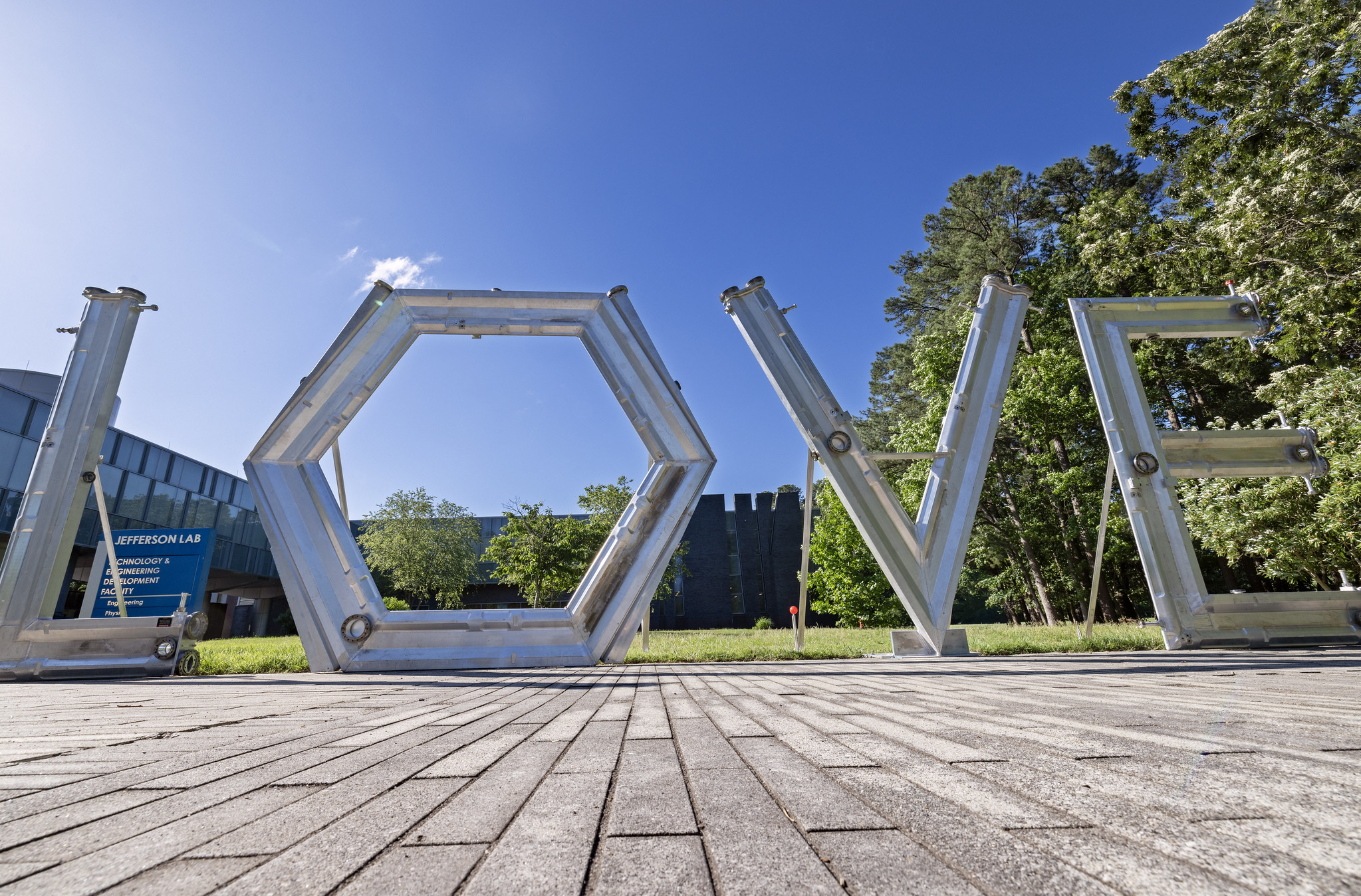 Giant LOVE letters are crafted from aluminum vacuum chambers along the edge of a brick walkway in front of a building with grass and trees in the background at Jefferson Lab.